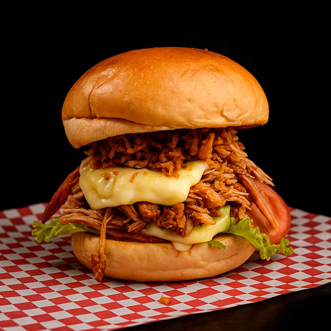 Hamburguesa BurgueChona en pan brioche con lechona desmechada, doble queso fundido y papas crocantes, servida sobre papel de cuadros rojos.