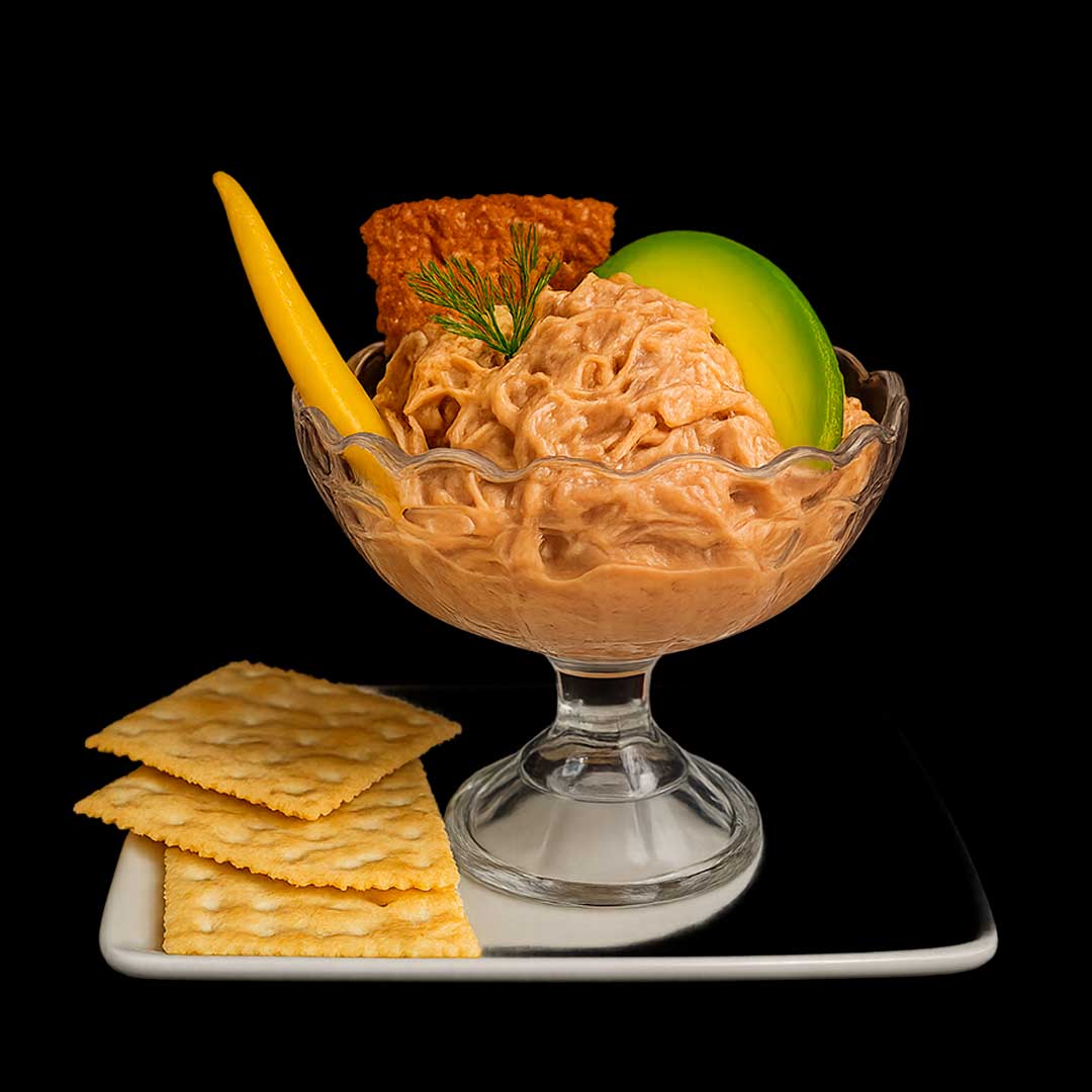 Copita de vidrio con ceviche de lechona cremoso, decorado con lámina de aguacate, bastón de chicharrón, ají dulce y acompañado de galletas de soda sobre plato blanco, fondo negro.