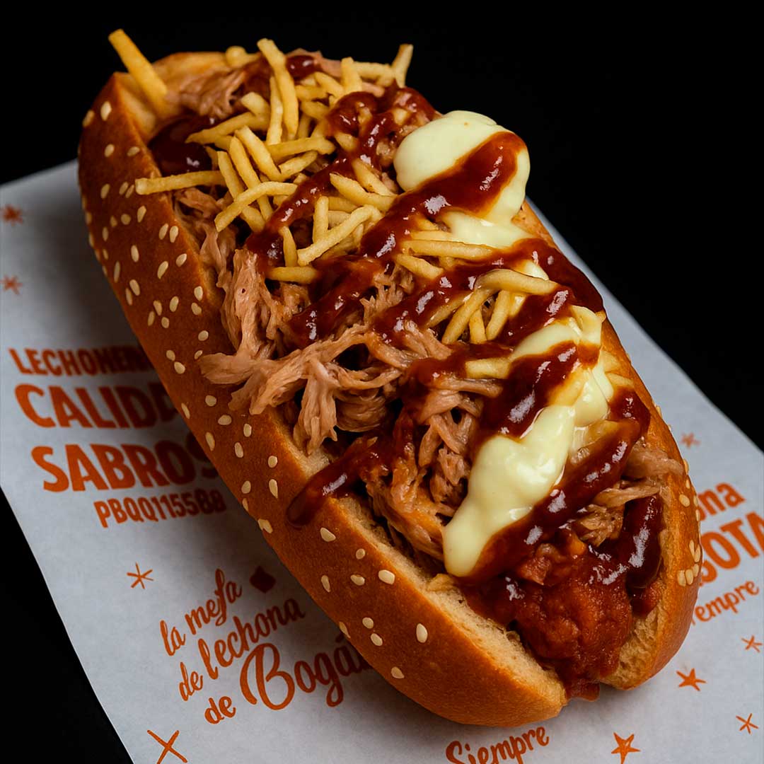 Hot dog de pan con ajonjolí relleno de lechona desmechada, papa chip, salsa BBQ y queso cremoso, sobre papel encerado con logo de la lechonería.